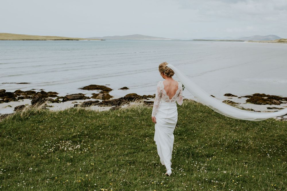 A bride in white dress walks along coastal grassland overlooking Scottish waters.