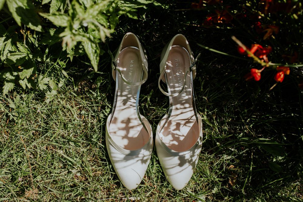 Elegant white and silver wedding shoes resting on grass with greenery in the background.
