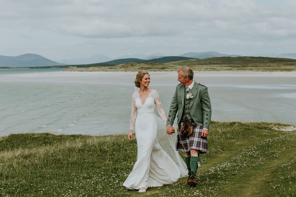 A couple walking along coastal grassland with white sand beach in background.
