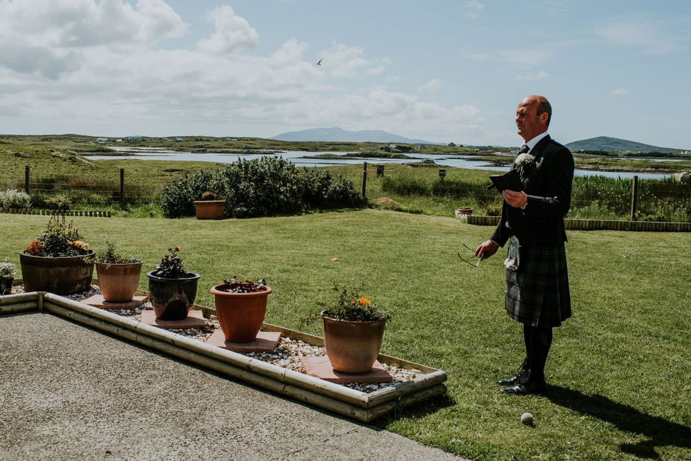 Man in traditional Scottish kilt stands by potted plants overlooking coastal landscape.