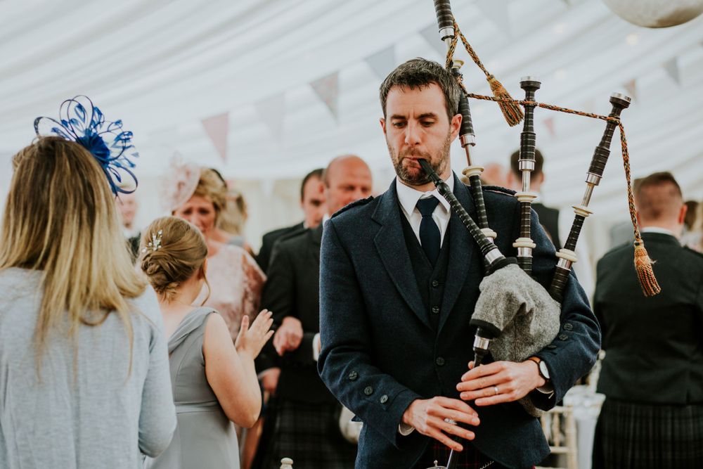 Bagpiper performing at an indoor wedding celebration with guests in traditional Scottish attire.