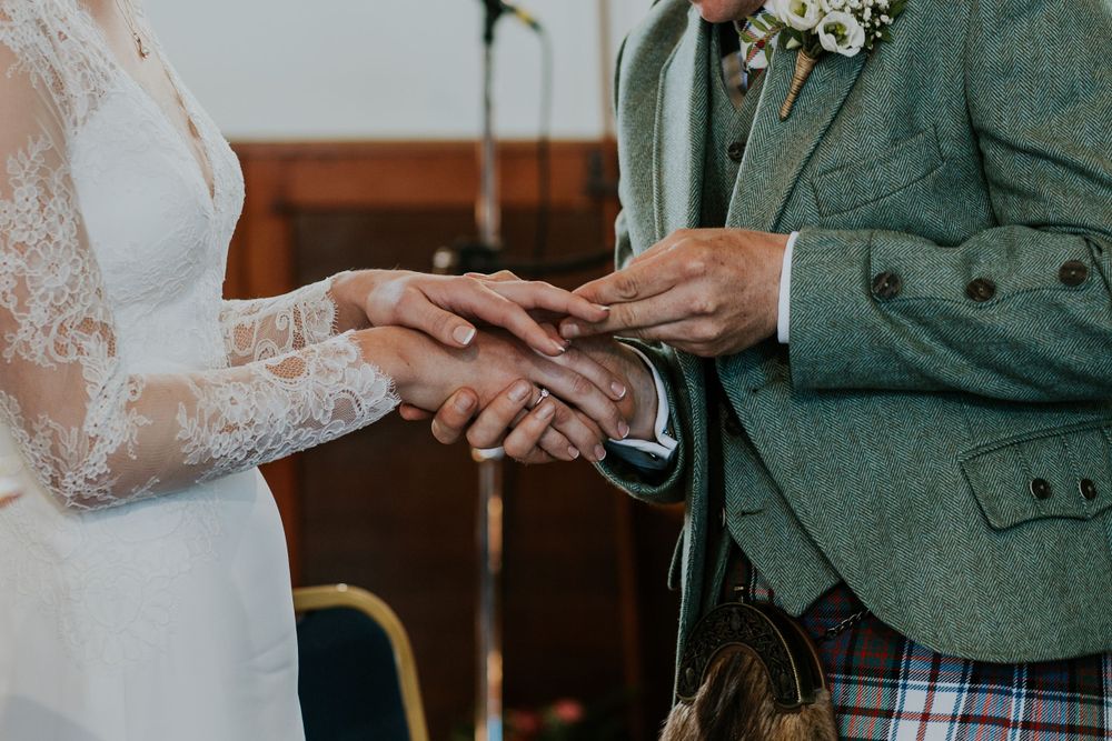 Close-up of wedding ring exchange with green Scottish jacket and white lace dress.