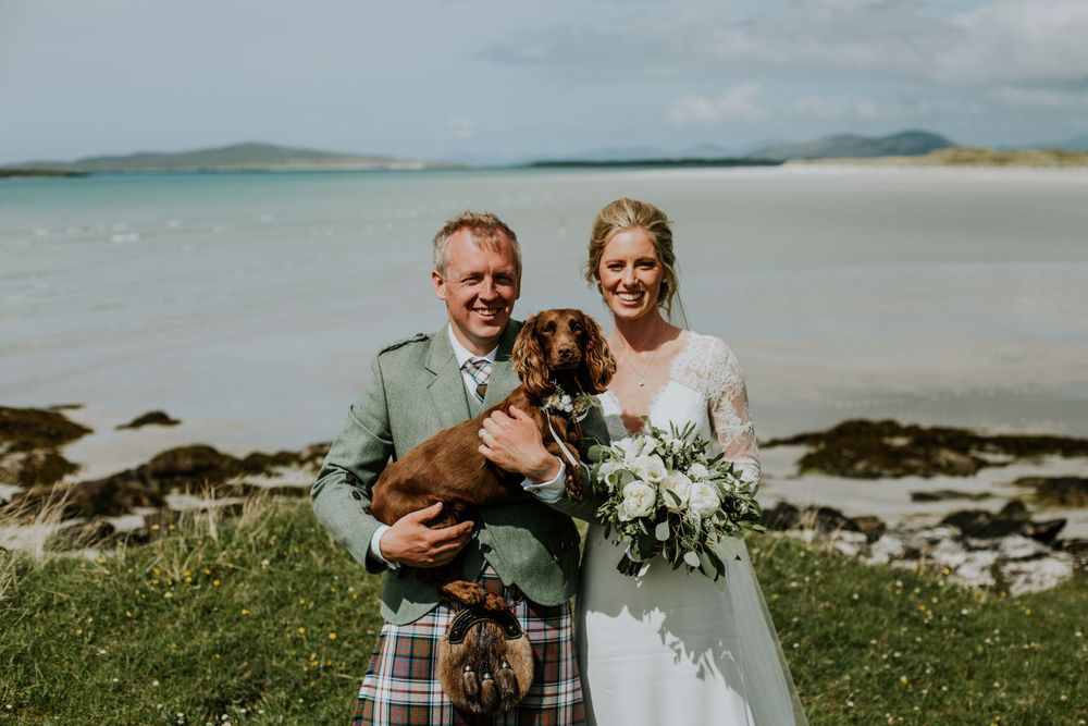 A wedding couple poses with their brown dog on a scenic beach with mountains in the background.