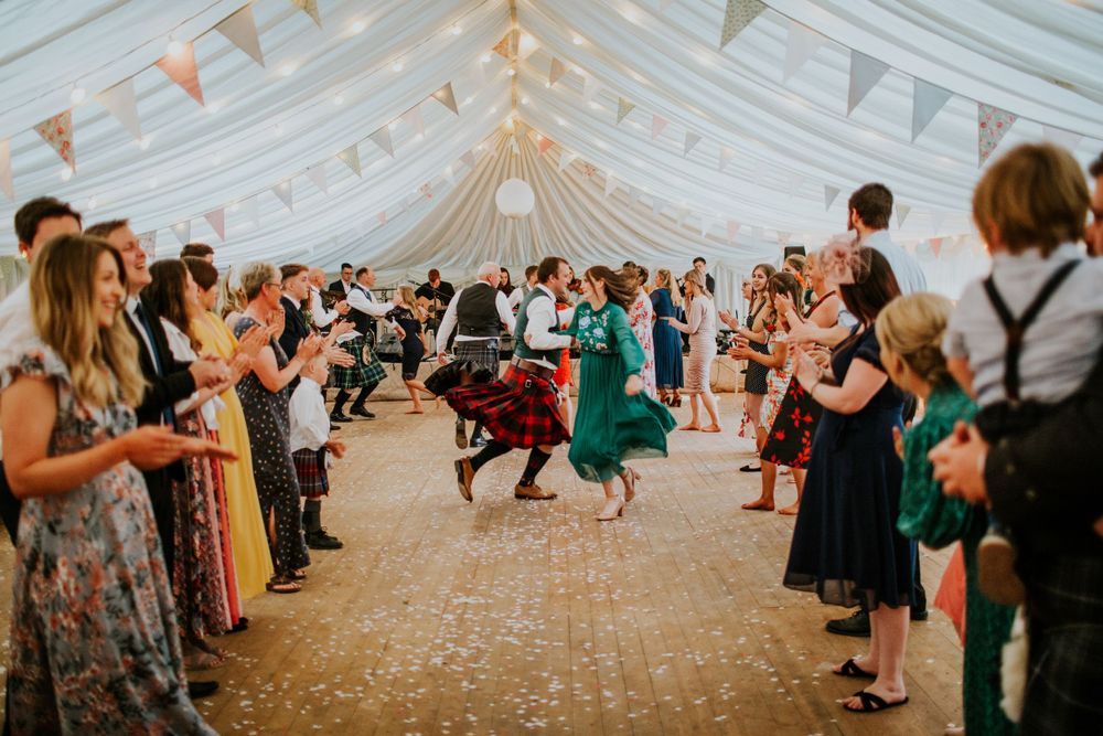 Wedding guests dance and celebrate inside a white tent decorated with bunting flags at a lively reception.