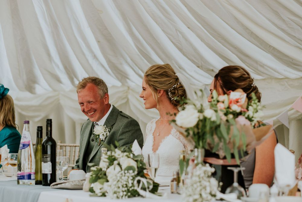 Wedding party seated at head table with floral centerpieces under white tent.