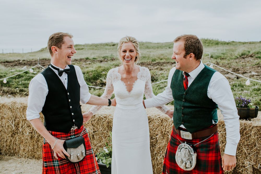 Wedding party in traditional Scottish kilts poses outdoors against rural backdrop.