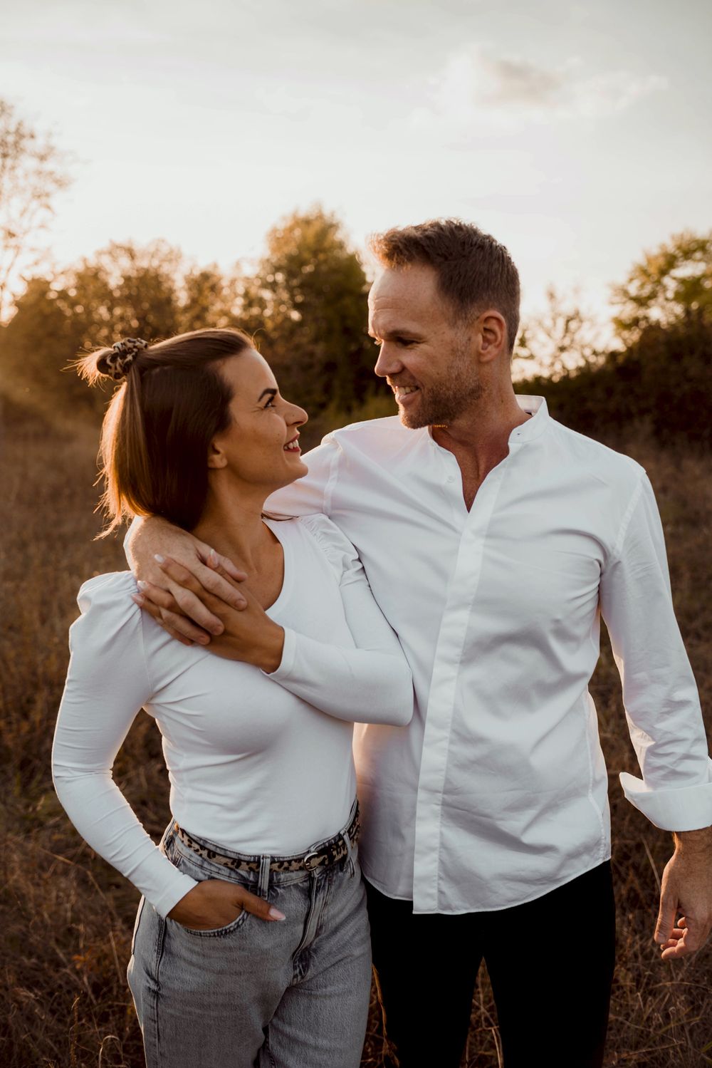 Couple shares loving glances while embracing in golden hour light among tall grass.