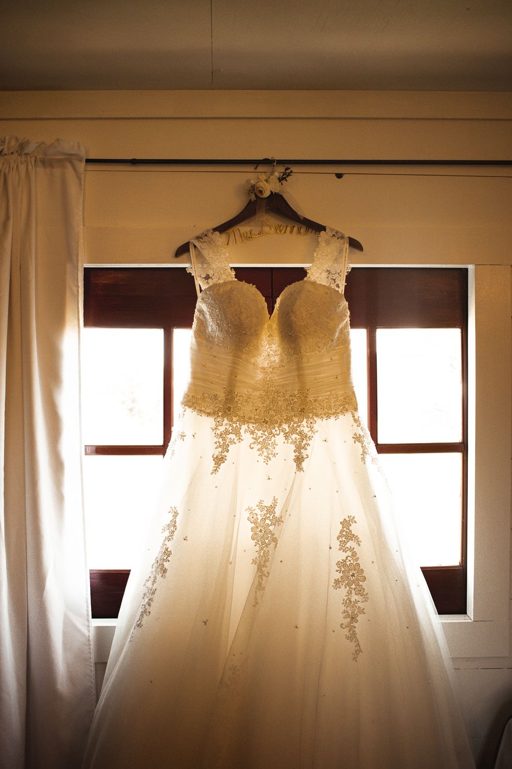 Elegant white lace wedding dress hanging in window light with delicate beaded details.