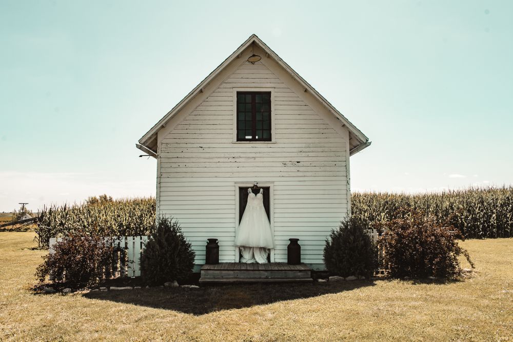 A stark white farmhouse stands alone among cornfields under a pale blue sky.