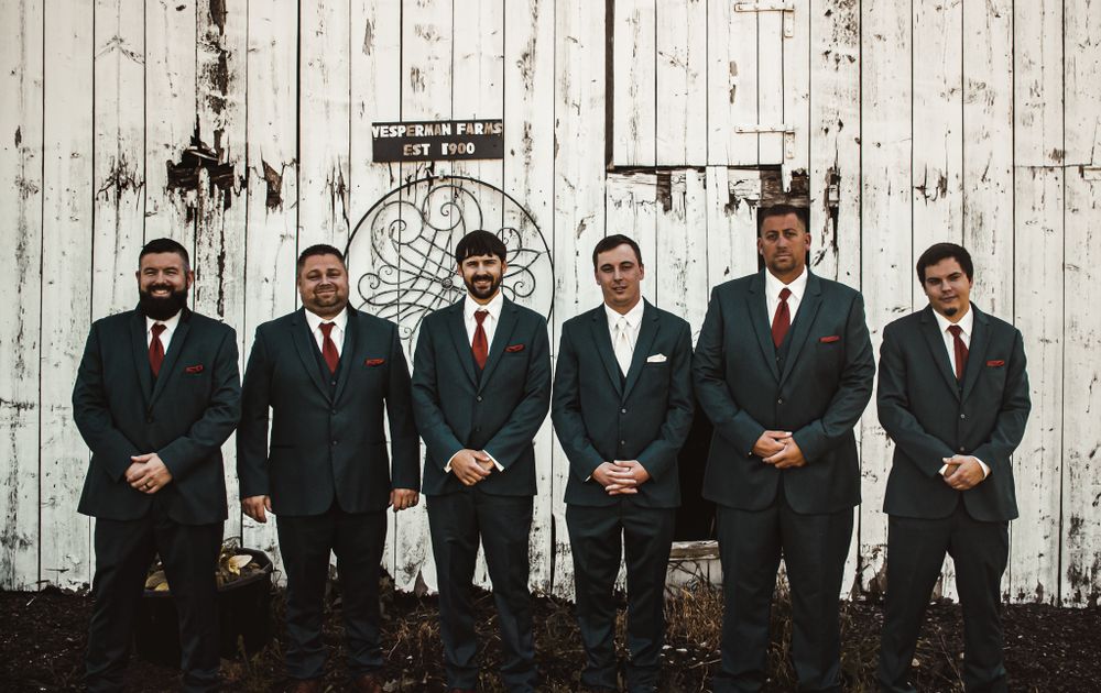 Groomsmen in dark suits pose together against a rustic white wooden wall backdrop.