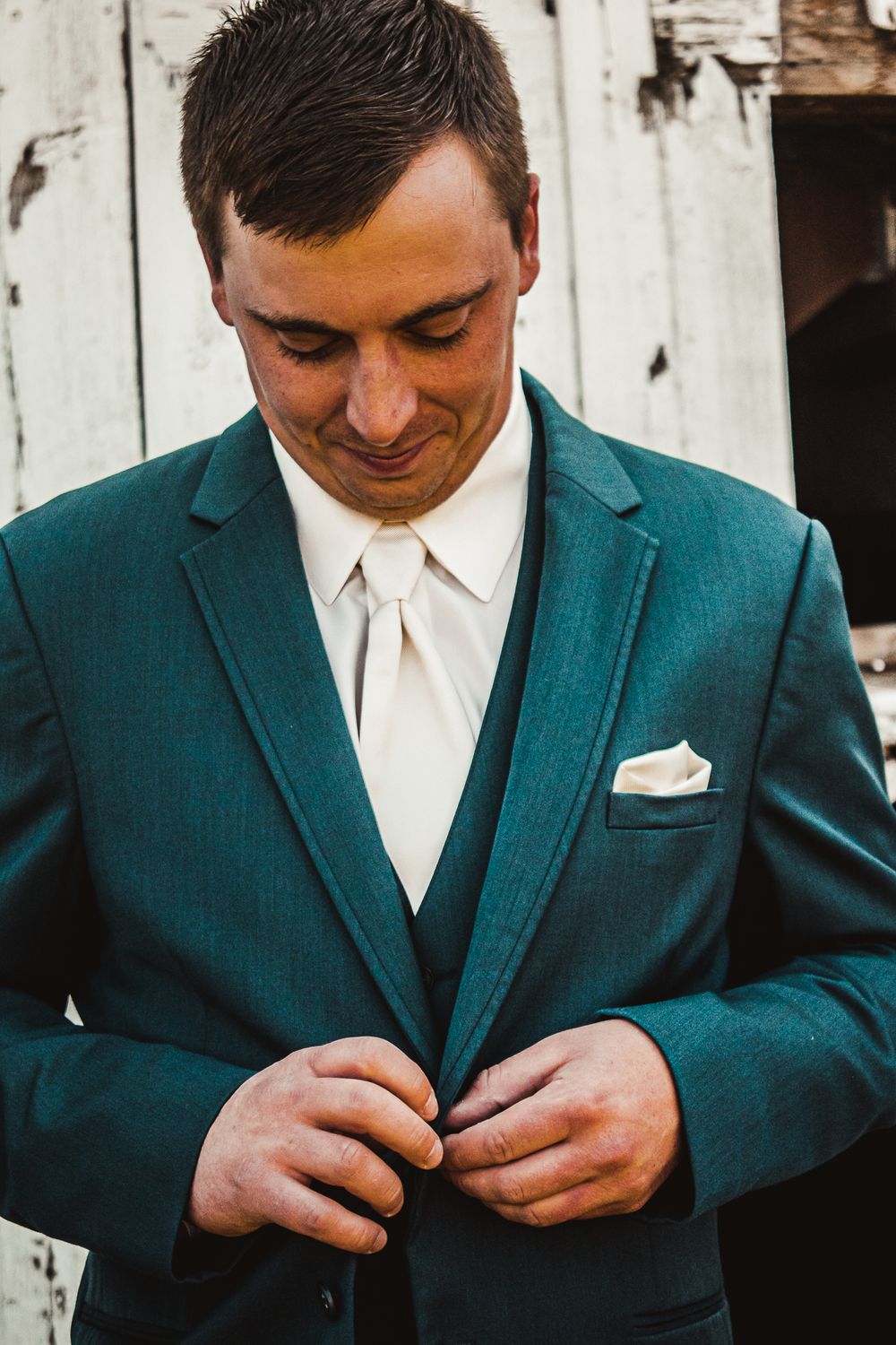 A person wearing a teal suit and white tie adjusts their buttons against a white wooden wall.