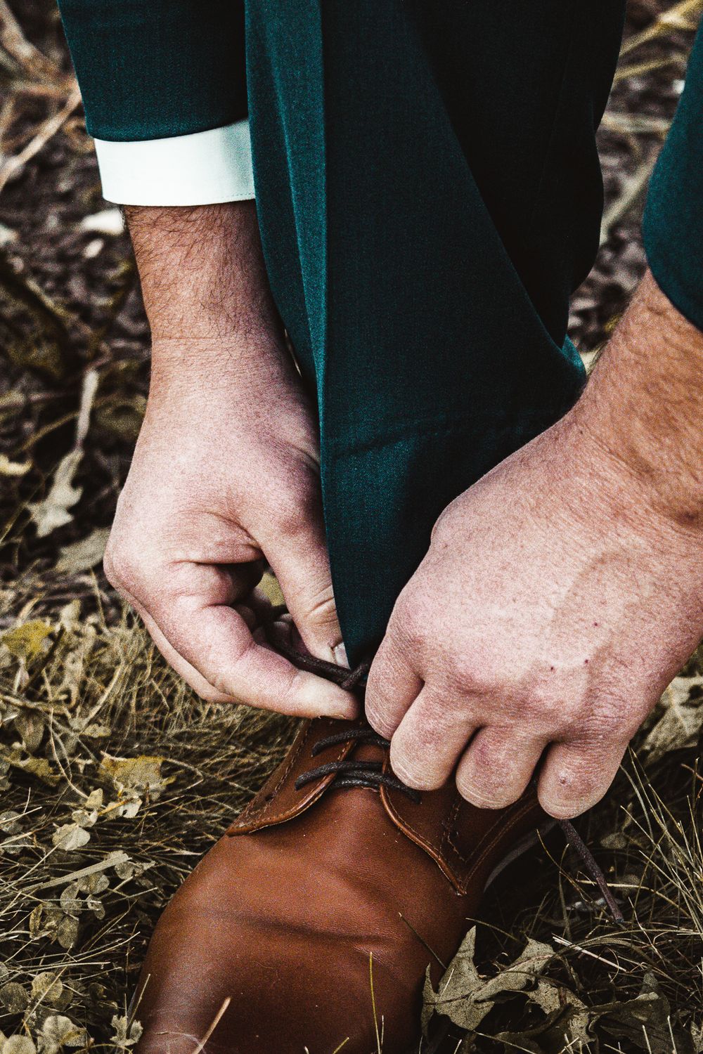 Close-up of hands tying brown leather dress shoes with teal pants visible.