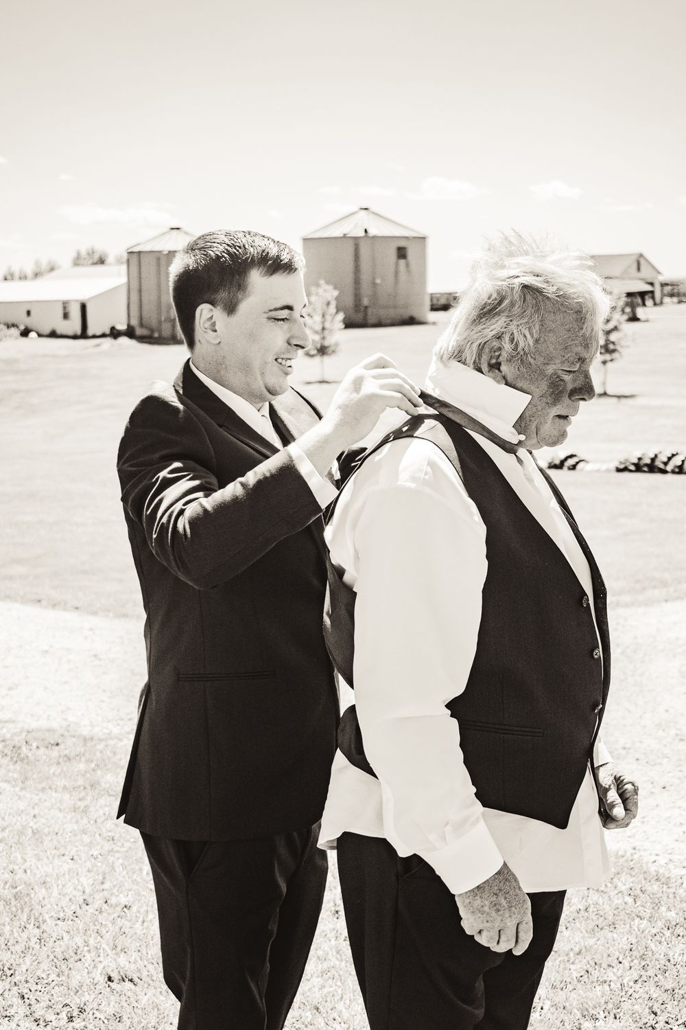 Black and white photo showing someone helping adjust another's formal attire outdoors.