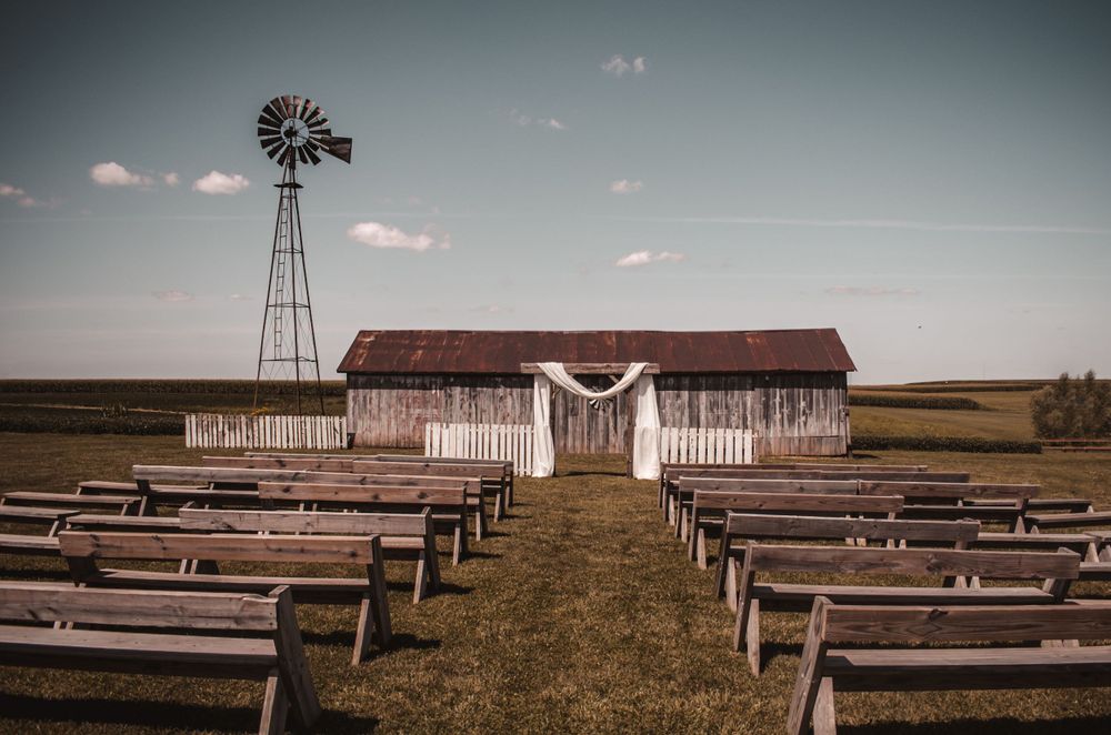 Rustic outdoor wedding venue with wooden benches facing a barn and windmill under a cloudy sky.