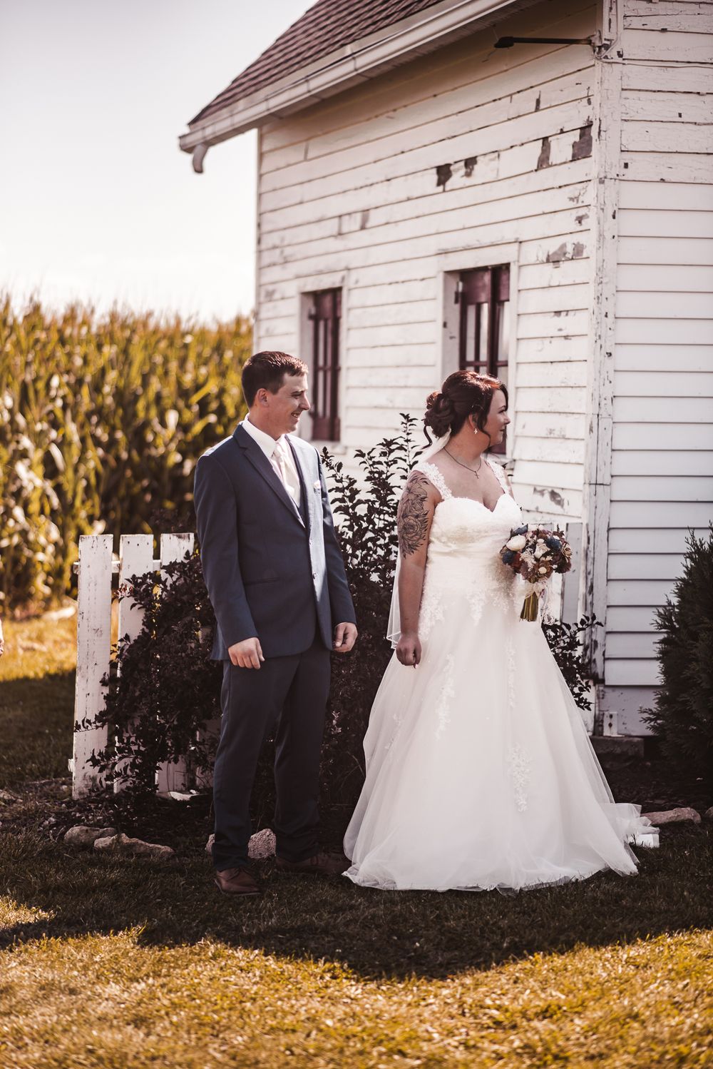 Newlyweds pose outside a white farmhouse next to a cornfield.
