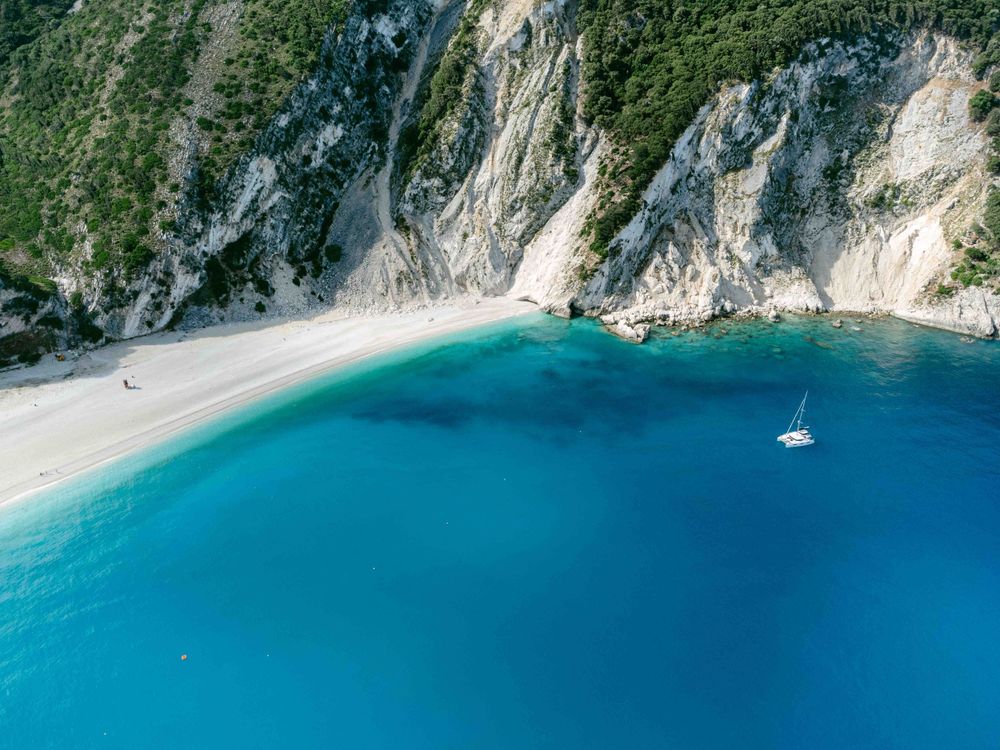Dramatic coastal cliffs meet crystal clear turquoise waters with a secluded beach and anchored yacht.