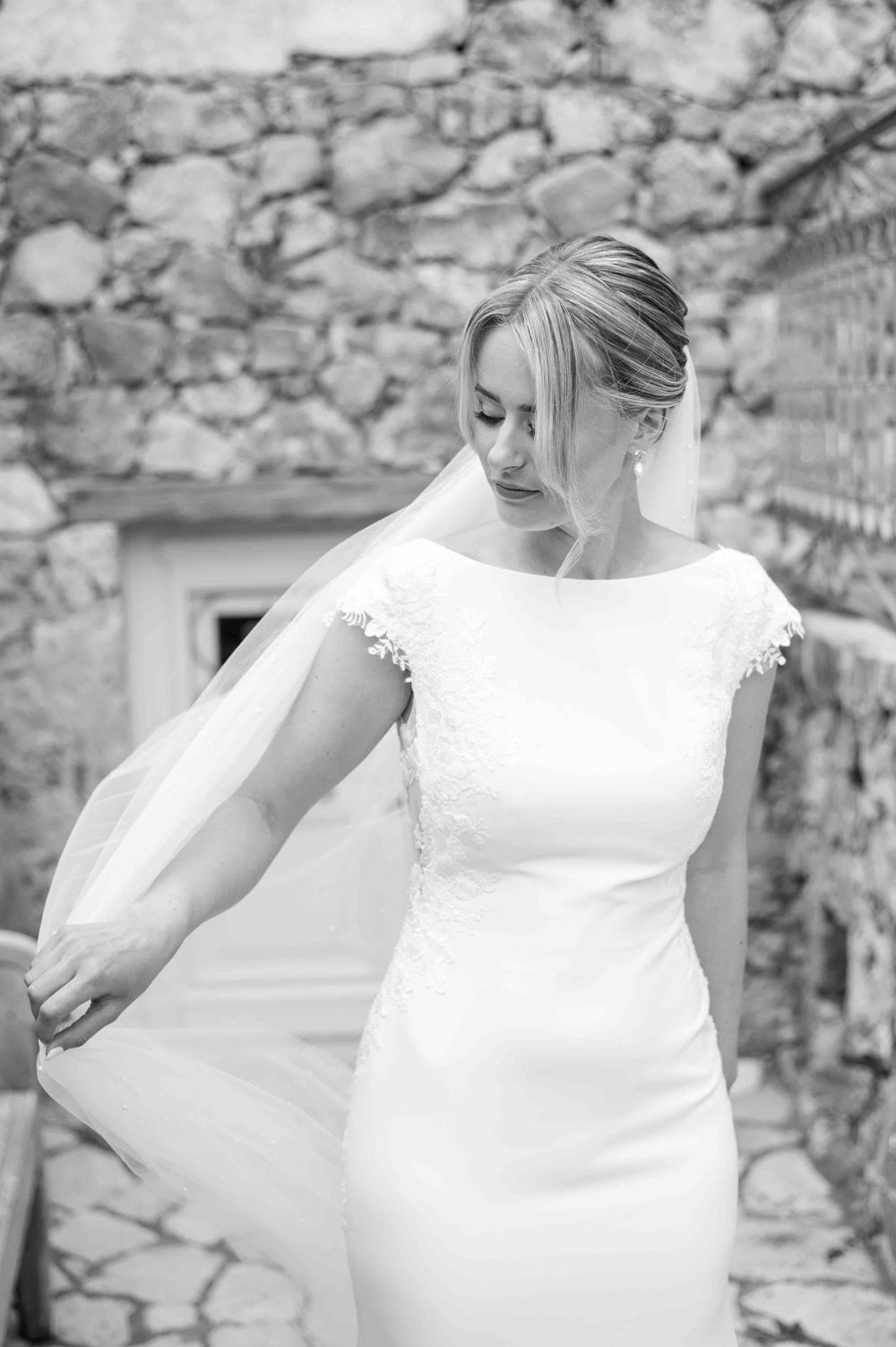 Elegant bride in a fitted white wedding dress adjusting her gown against a stone wall.