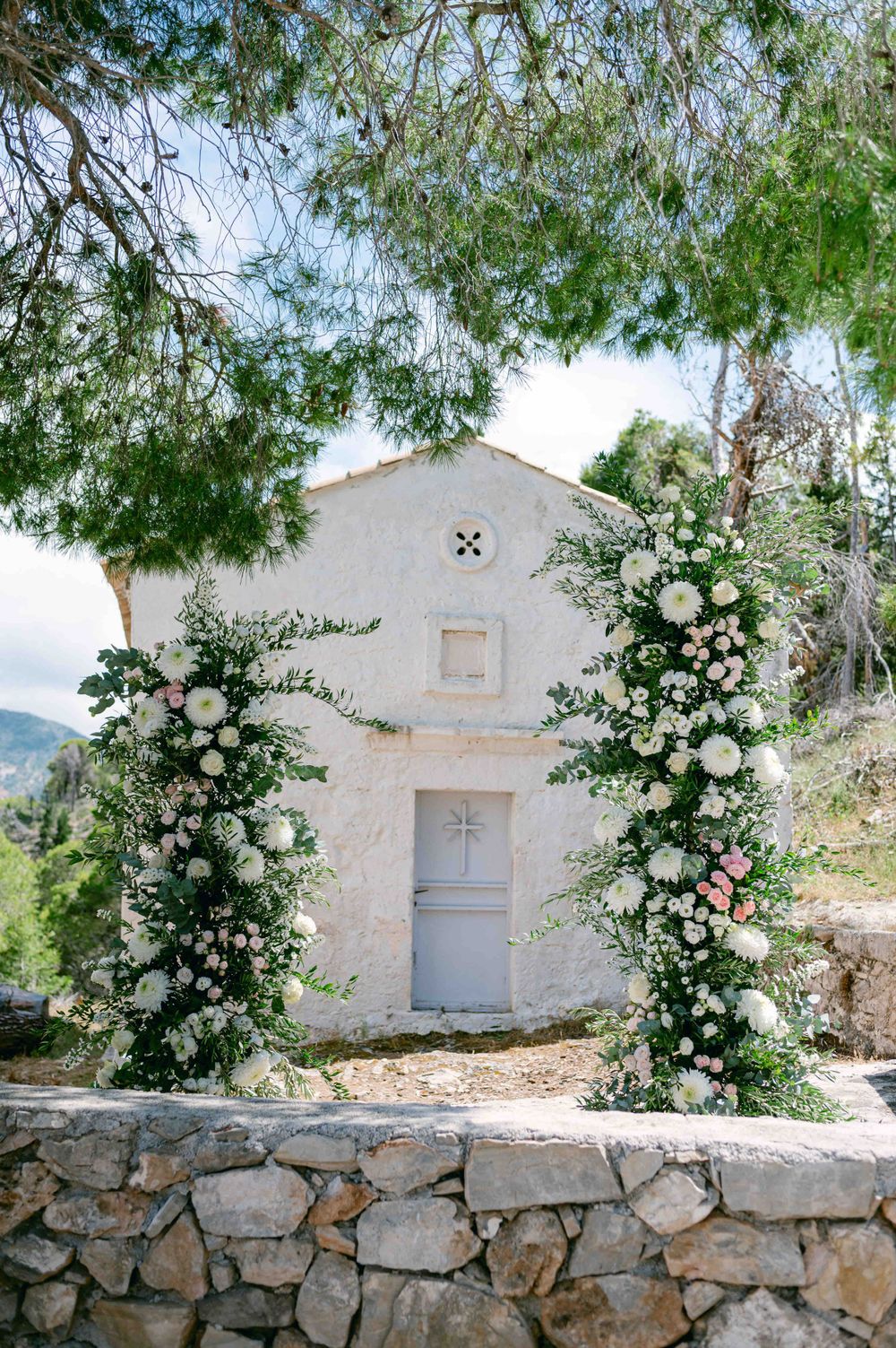 Small white chapel with floral archway featuring white roses and greenery against a mountainous backdrop.