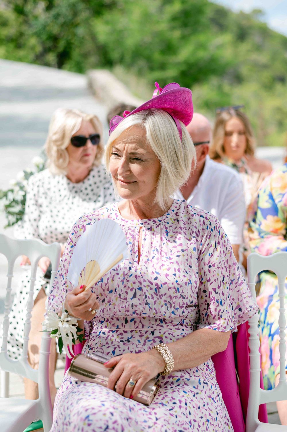 Wedding guest in floral dress and pink fascinator reading program at outdoor ceremony.