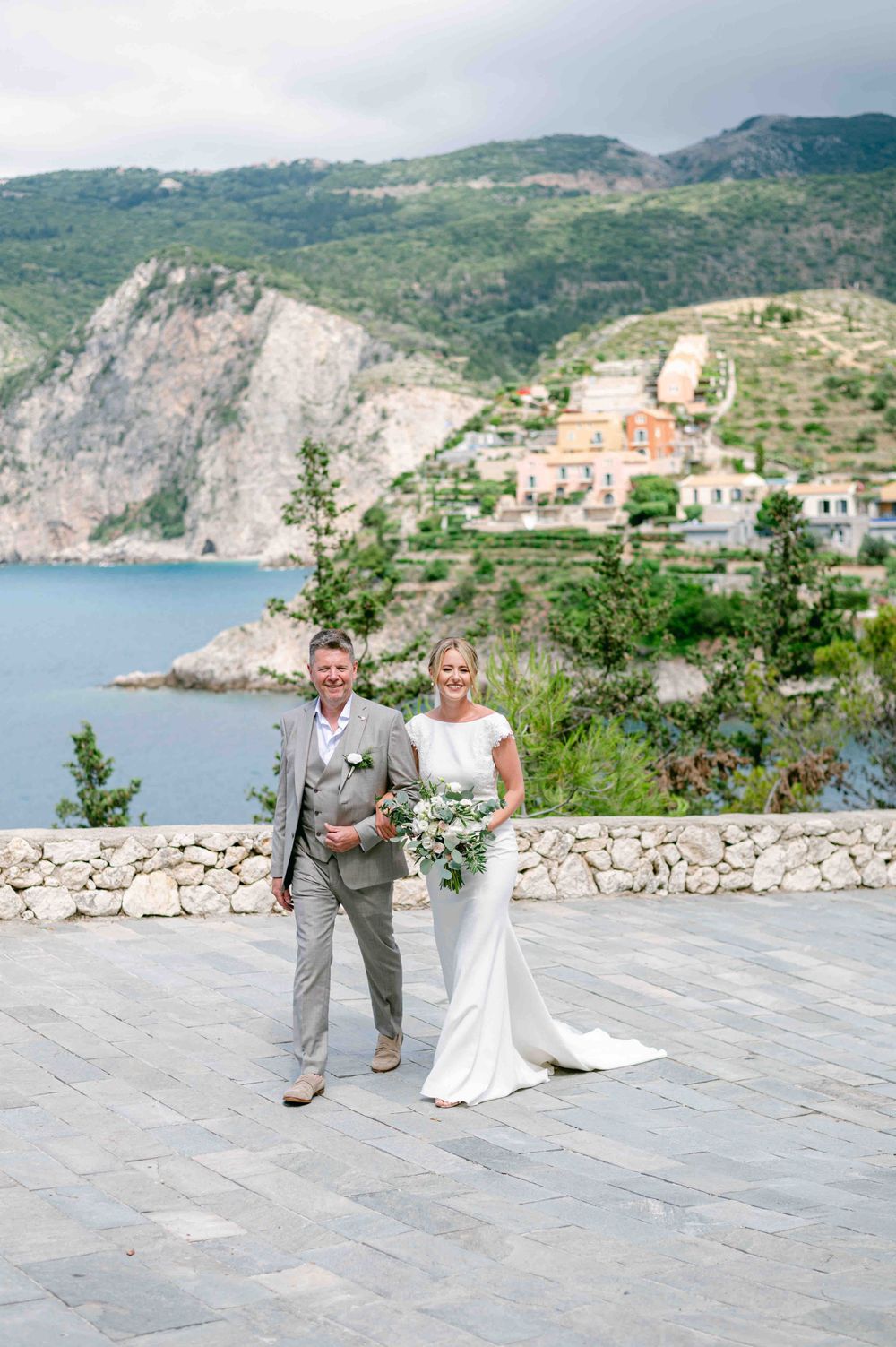 Elegant wedding couple poses on stone terrace overlooking Mediterranean coastline.