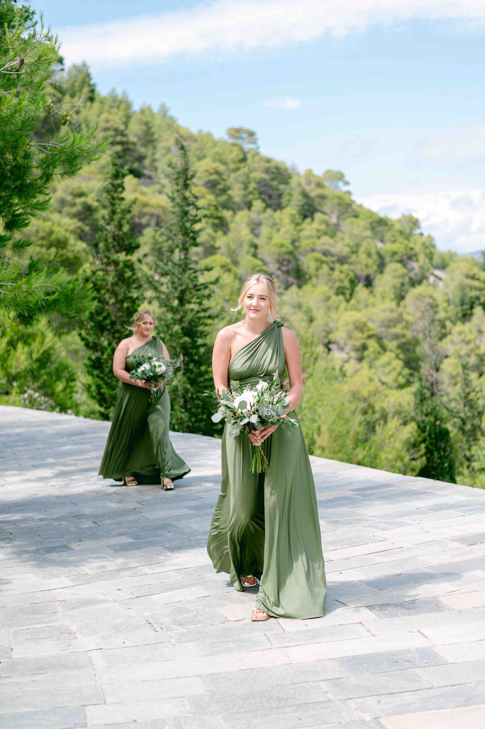 Bridesmaids in flowing sage green dresses walking on stone terrace with mountain views.