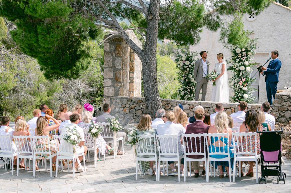 Outdoor wedding ceremony with white chairs and floral arch against mountain backdrop.
