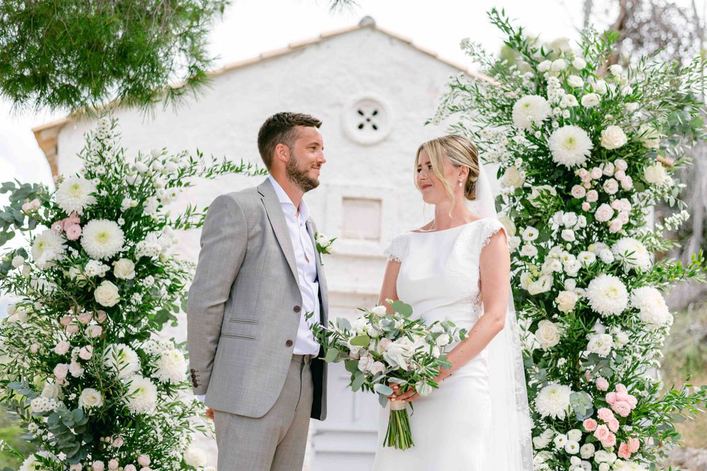 Wedding couple standing between white floral arches in front of charming chapel.