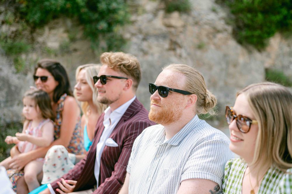 Wedding guests seated outdoors smiling and wearing sunglasses on a sunny summer day.