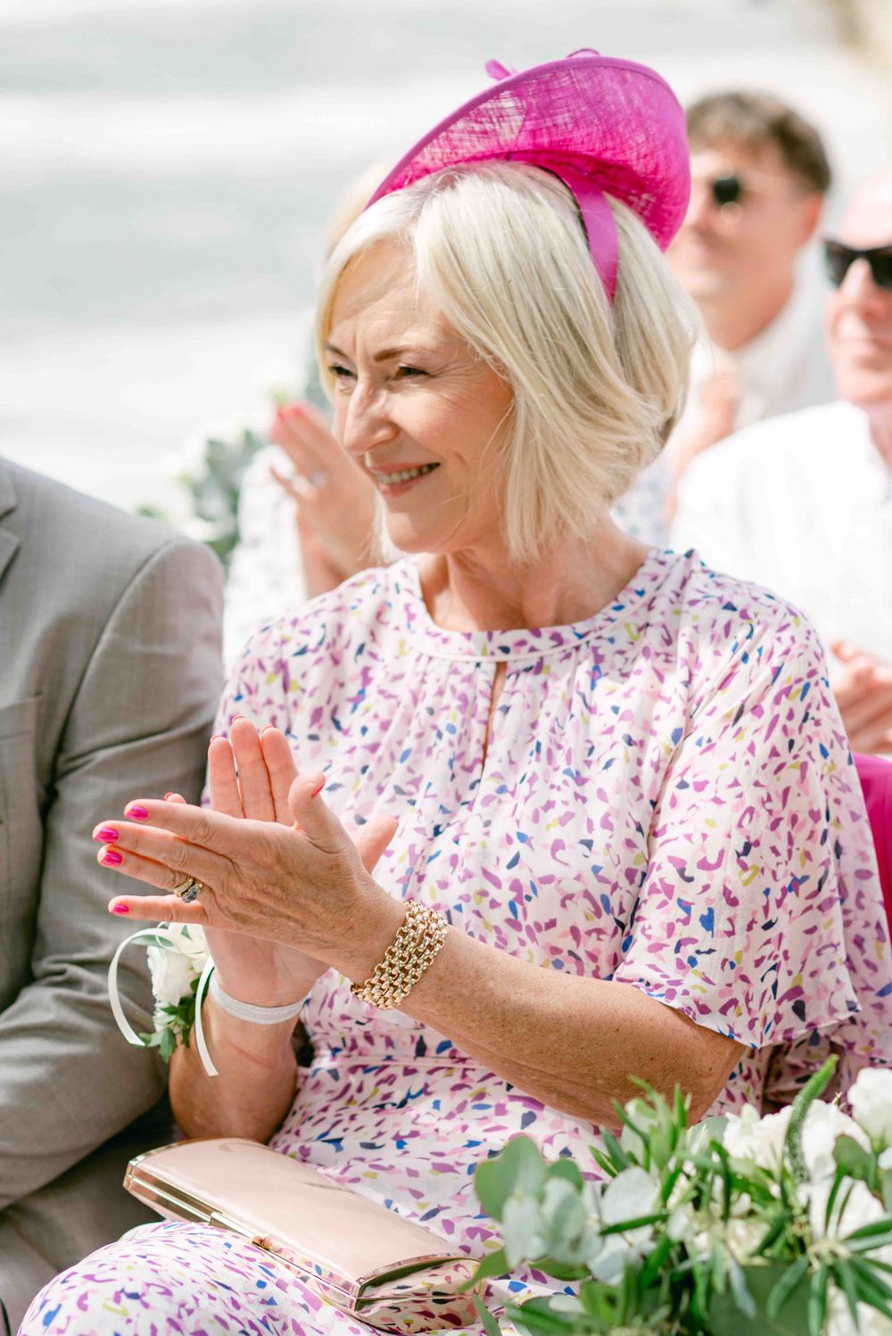 Guest in pink floral dress and matching fascinator smiling and clapping at outdoor event.