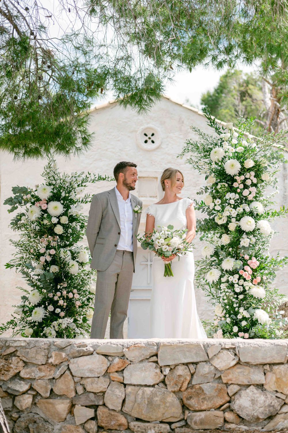 Couple standing under beautiful white floral arch against stone wall at outdoor wedding.