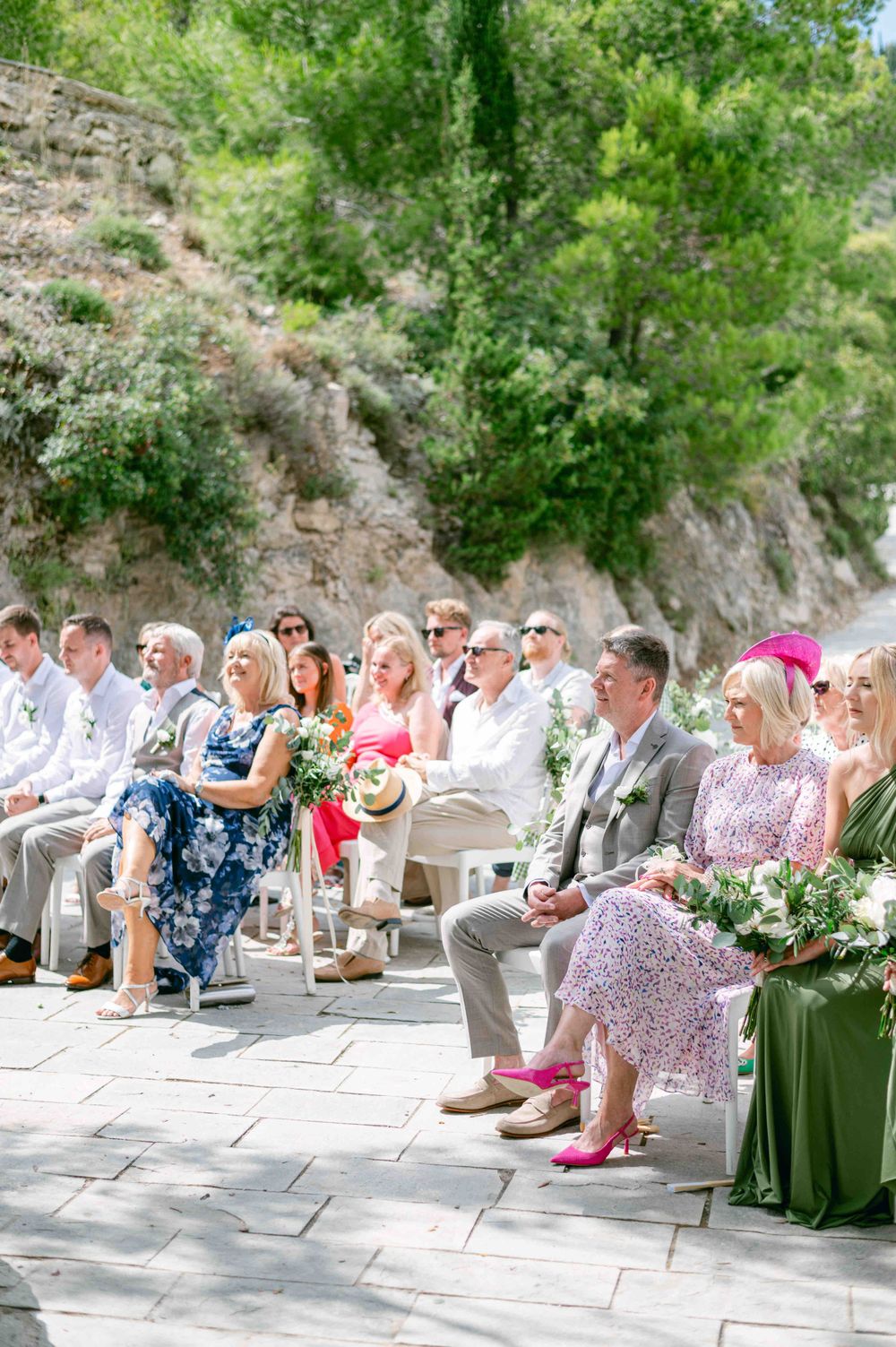 Wedding guests seated in white chairs at an outdoor ceremony with rocky cliff backdrop.