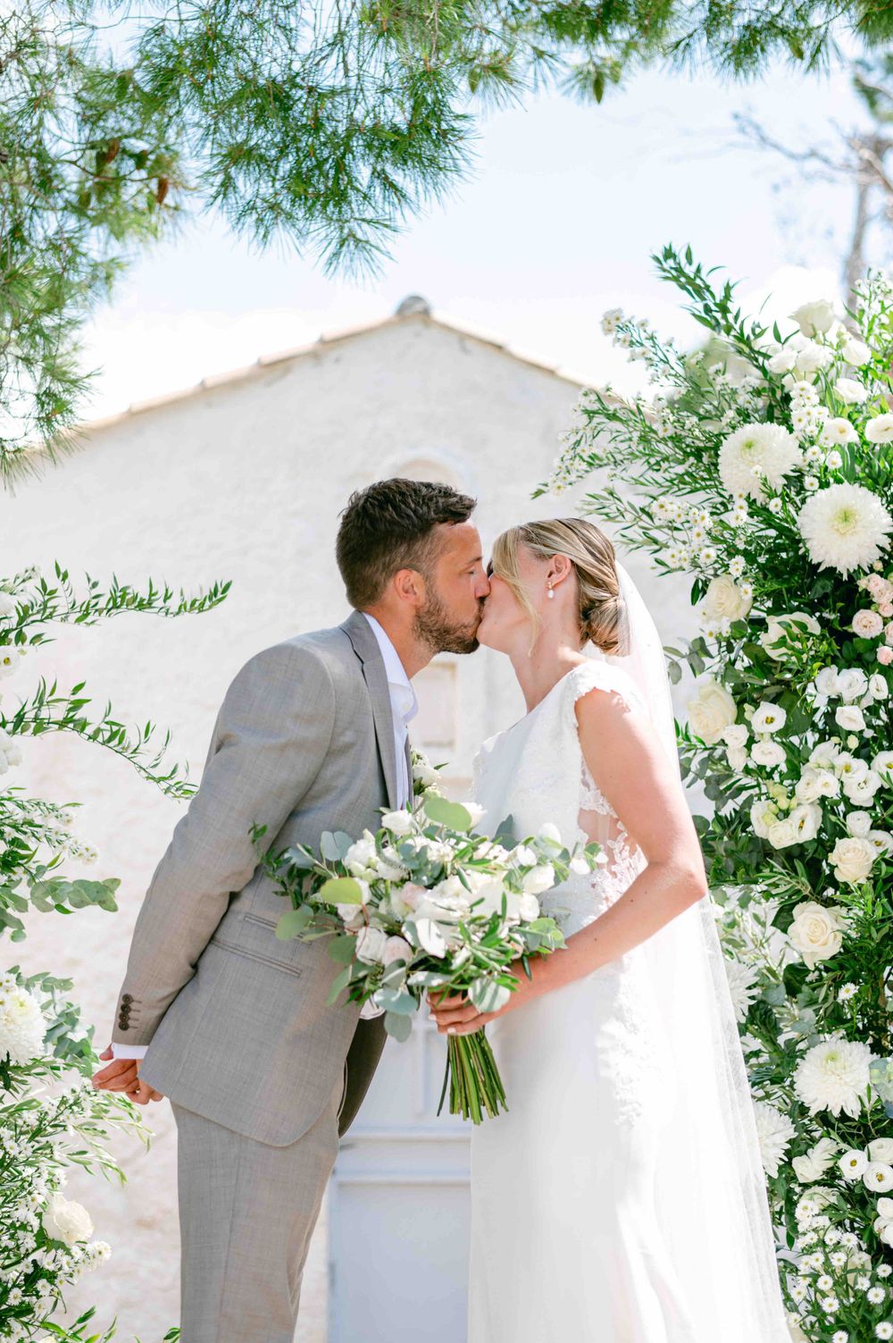 Newlyweds sharing first kiss under floral arch with white building in background.
