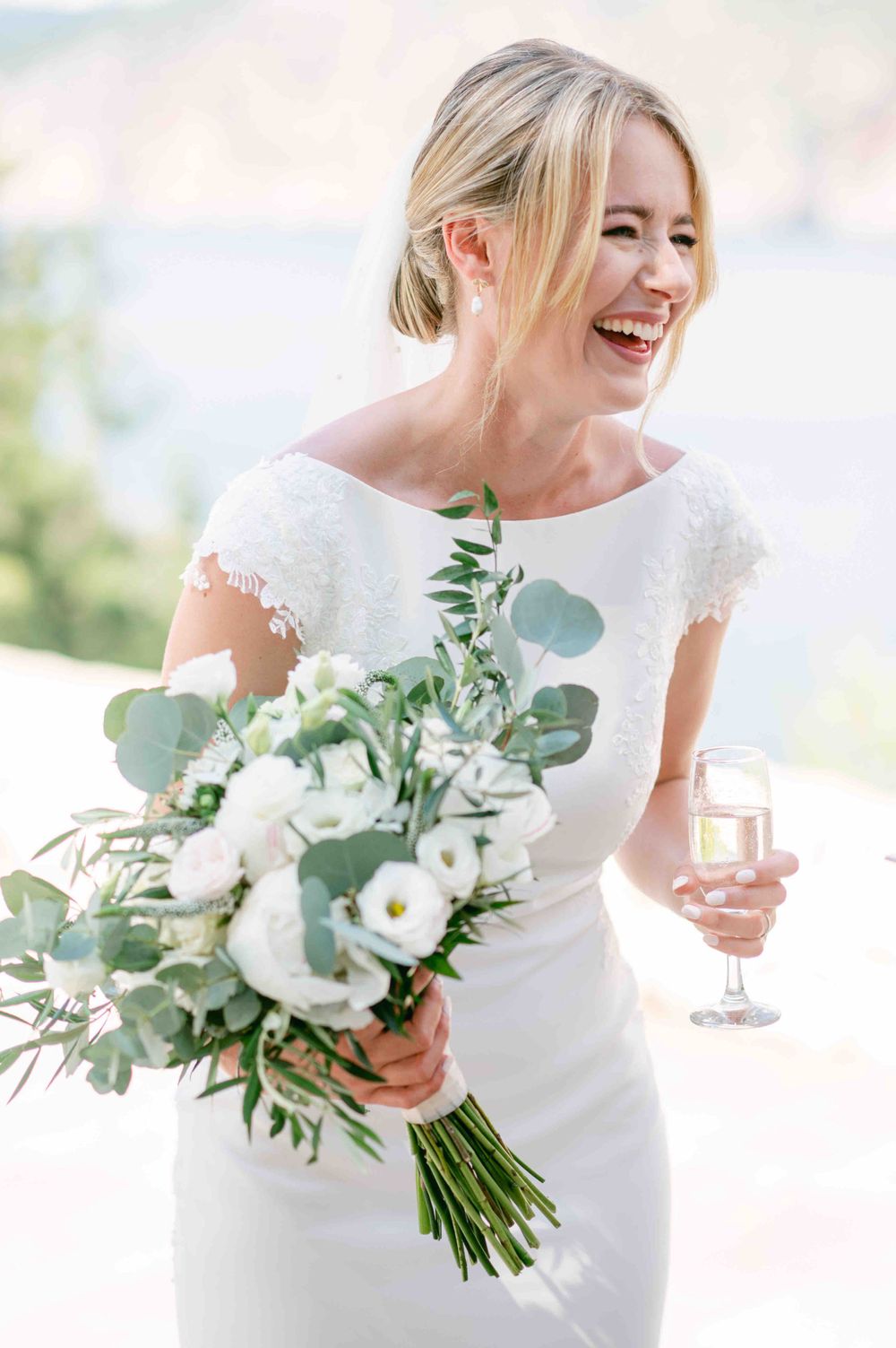 Joyful bride in classic white dress laughing while holding a lush eucalyptus and white flower bouquet.