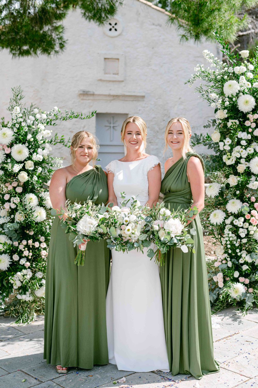Wedding party in white and sage green dresses posing against floral arch and white stone wall.