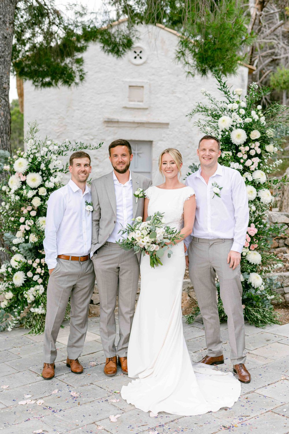 Wedding group photo with bridal party in light grey suits and white dress against floral backdrop.