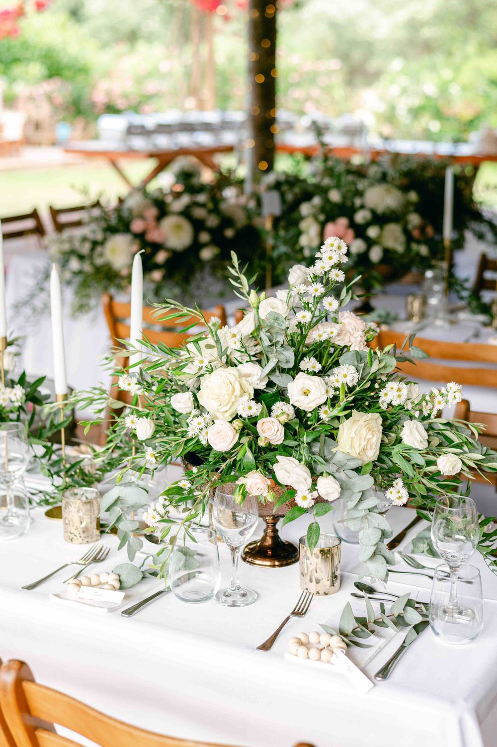 An elegant white floral centerpiece arrangement with roses and greenery on a formal table setting at an outdoor wedding reception.