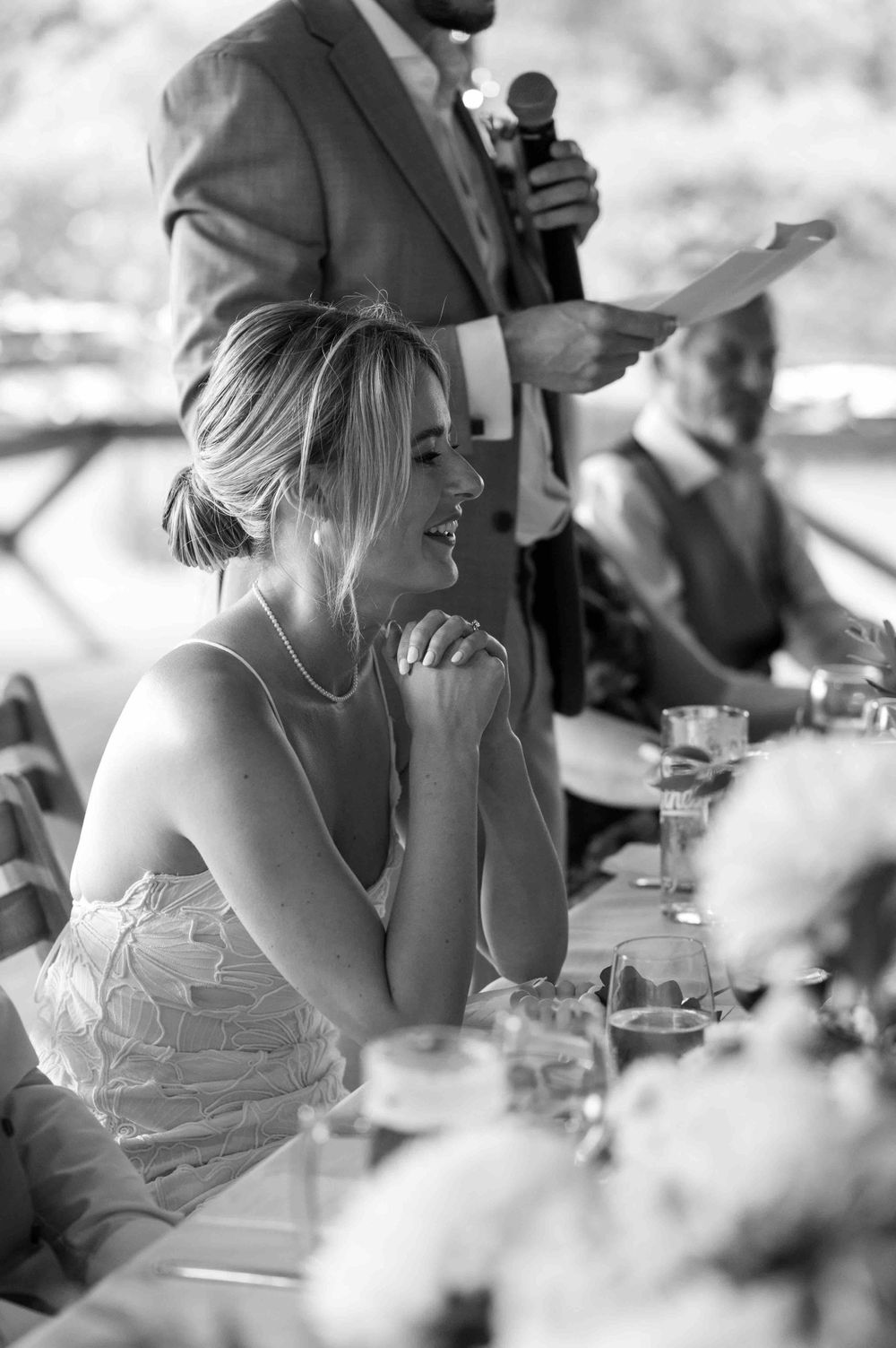 A candid black and white photo of a wedding reception speech moment showing guests seated at outdoor tables.