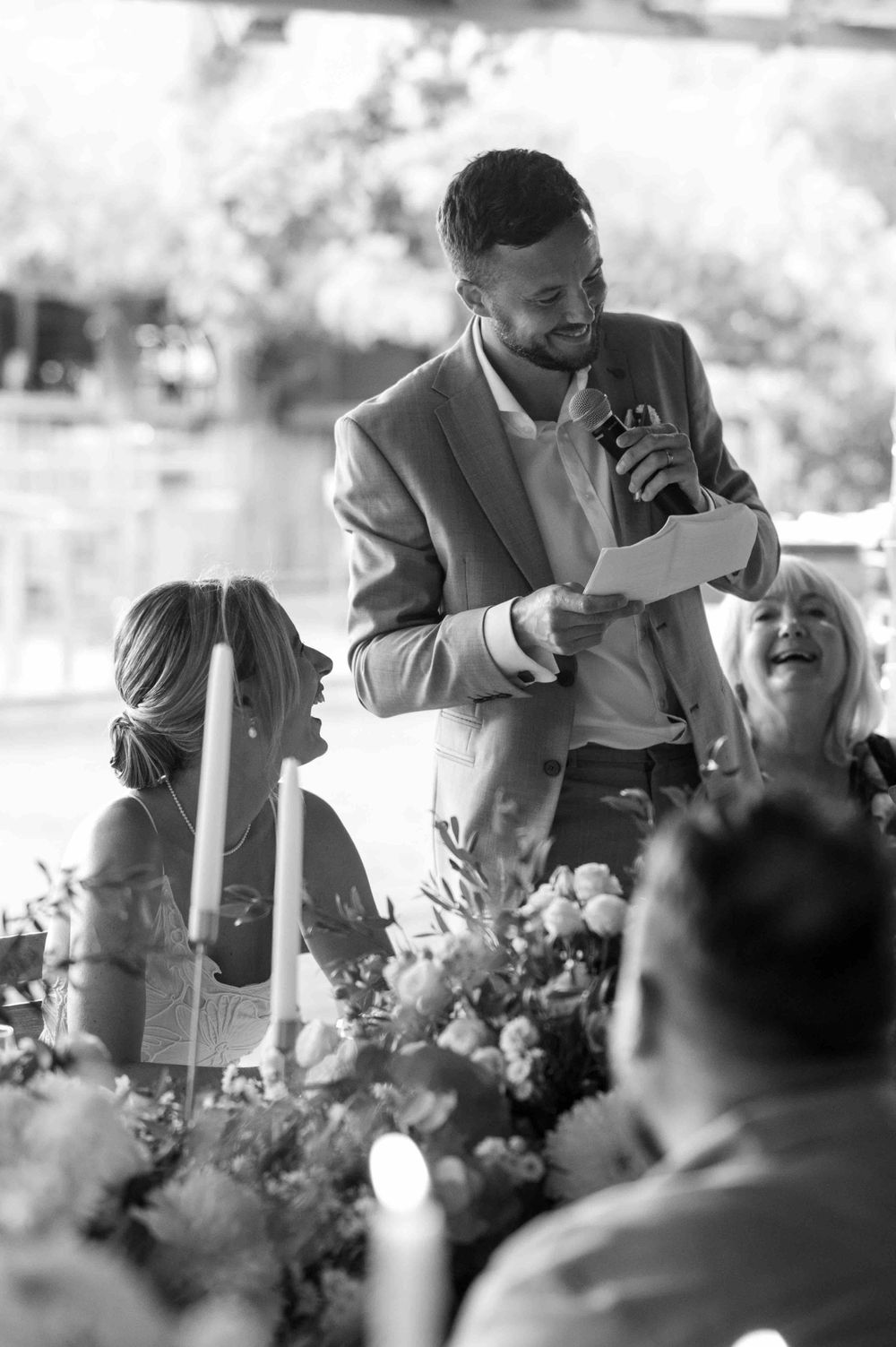 Black and white photo of someone giving a speech at an outdoor wedding reception with decorative floral arrangements.