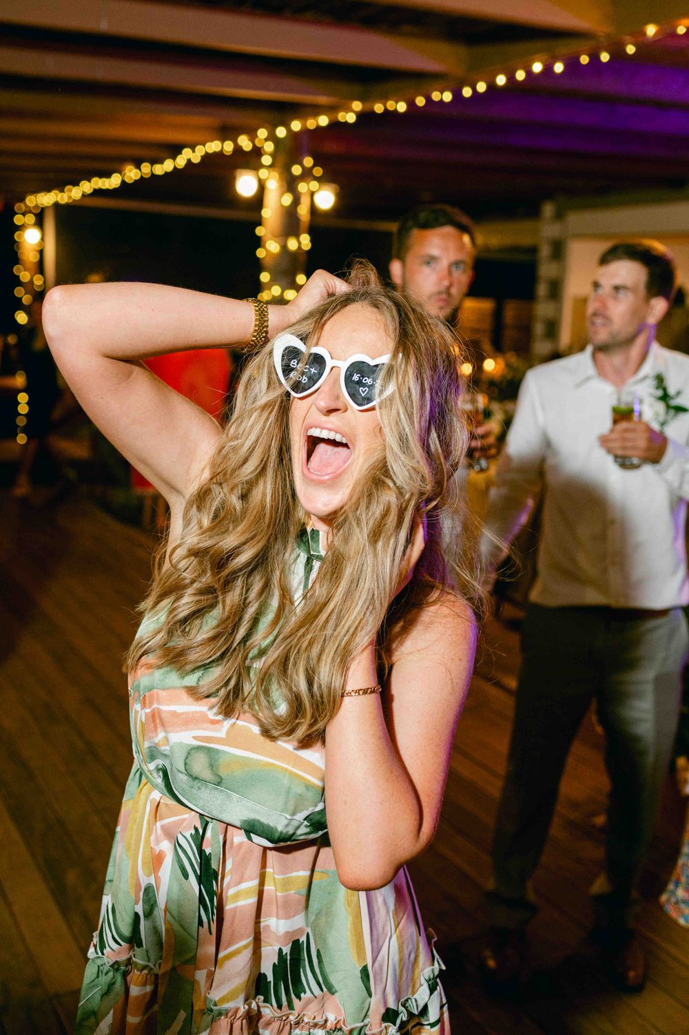 A joyful party guest wearing heart-shaped sunglasses dances under string lights at an evening celebration.