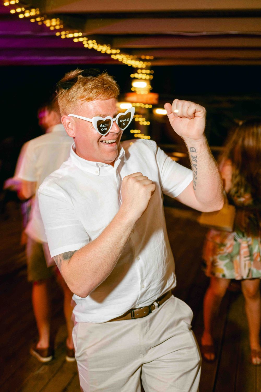 A guest wearing heart-shaped sunglasses dances enthusiastically under string lights at an evening party.