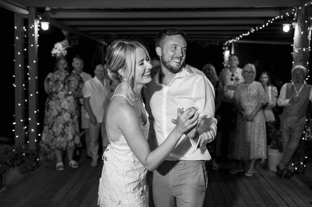 A black and white photo showing guests dancing and celebrating at a wedding reception under string lights.