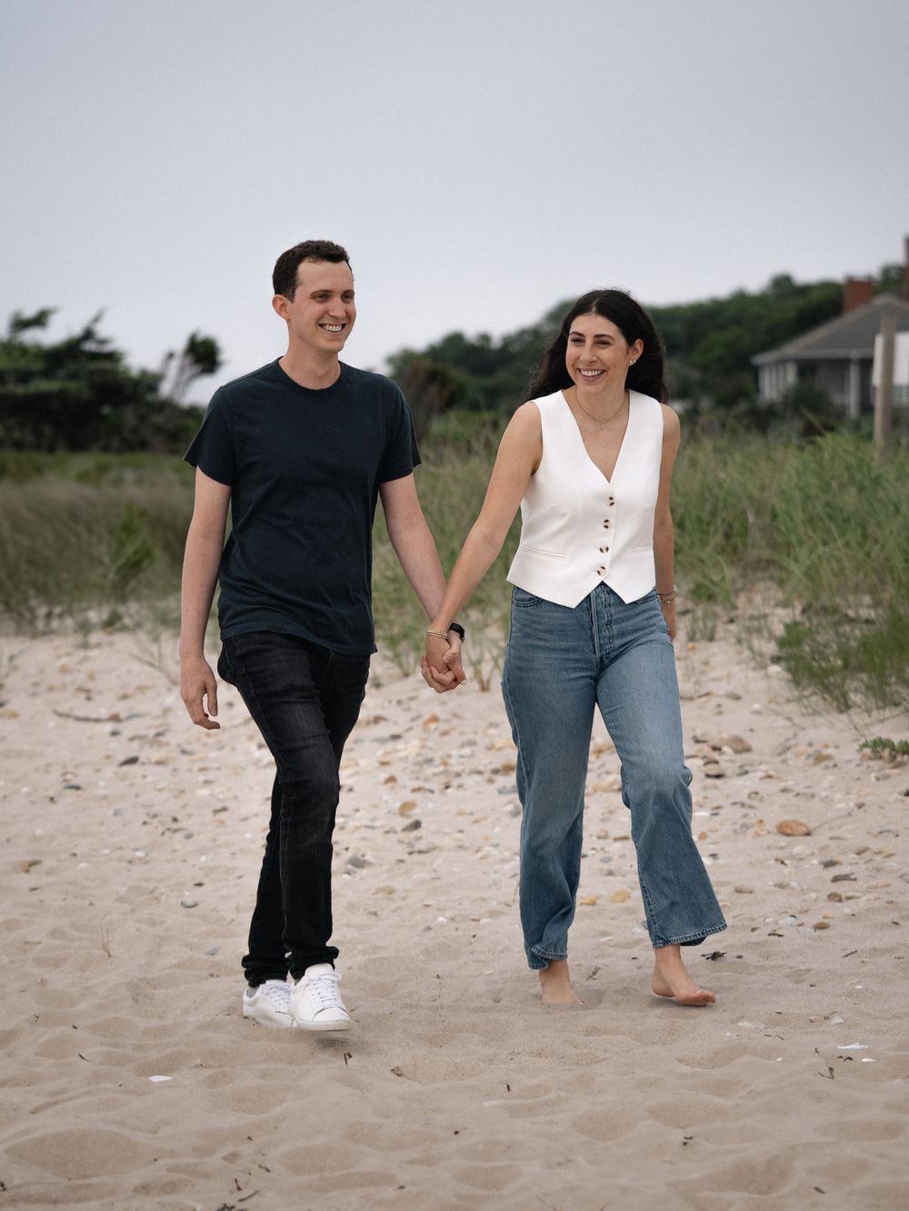 A couple strolls together barefoot on a beach path near coastal homes.