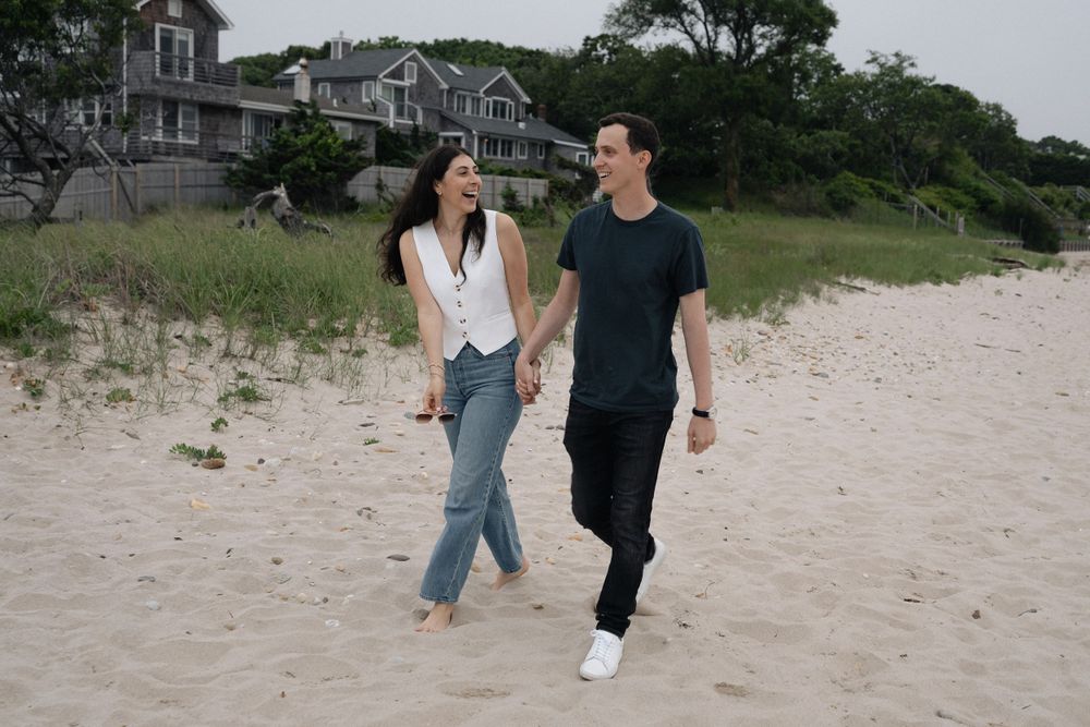 Two people walking barefoot along a sandy beach path near seaside houses.
