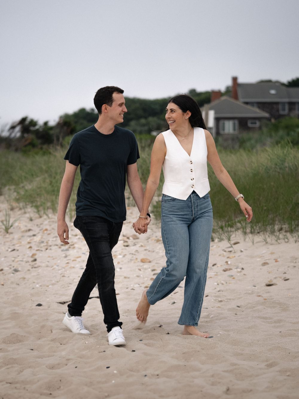 A couple walks hand in hand along a sandy beach path on a cloudy day.