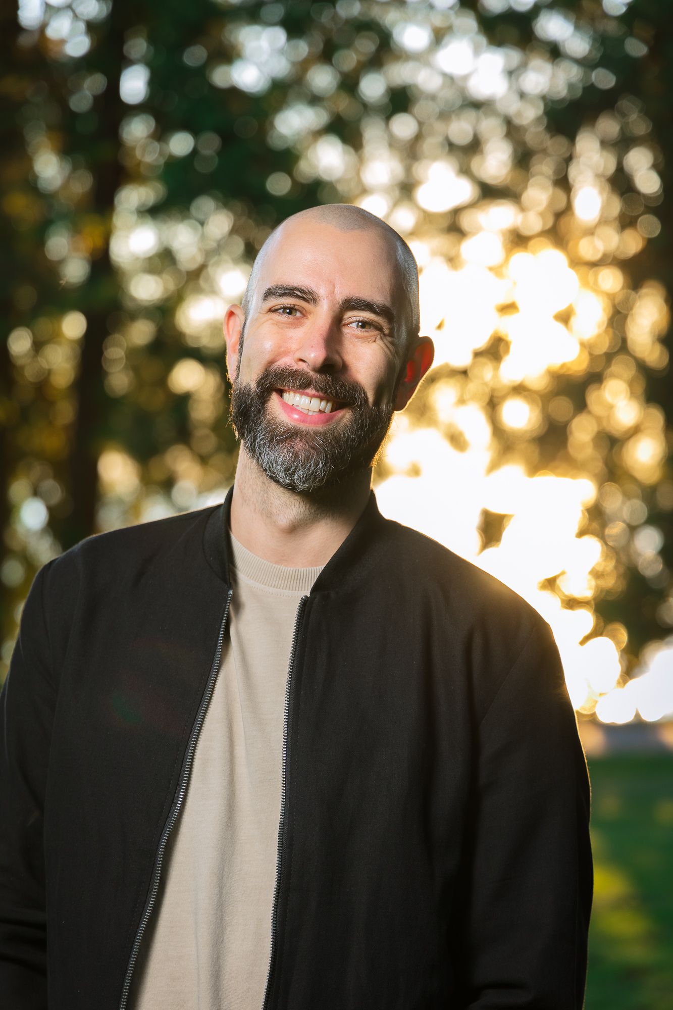 A bald, handsome man smiles wide for the camera. The sun sets in the trees behind him.