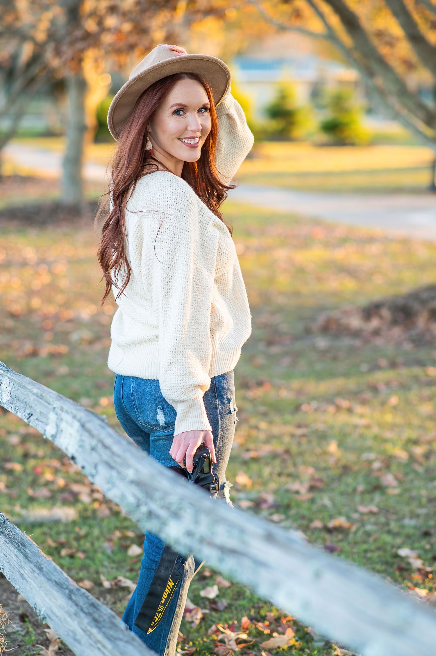 A woman in a white sweater and tan hat poses by a wooden fence on an autumn day.