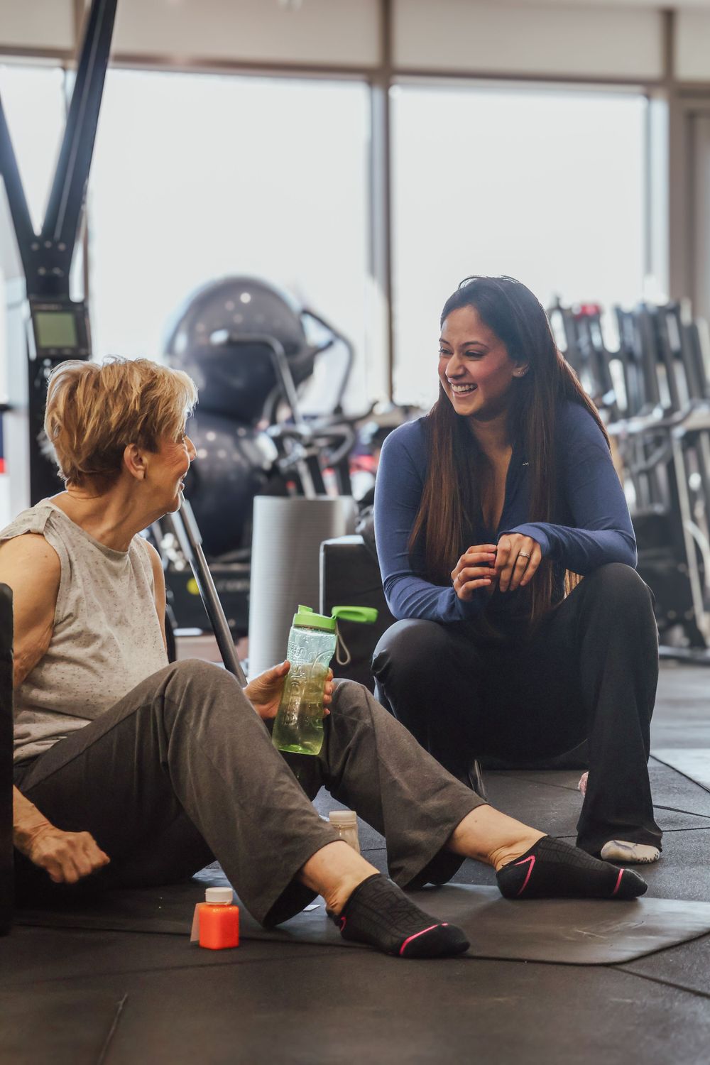 Two women sitting on a gym floor smiling and chatting during a break from their workout session.