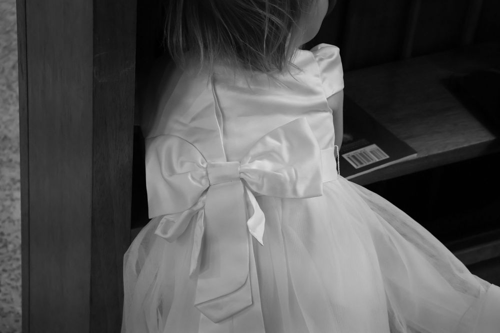 Black and white photo of a young girl in a white dress with a large satin bow on the back.
