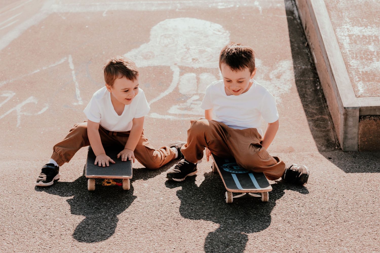 Two kids sitting on skateboards on concrete ground with chalk drawings.
