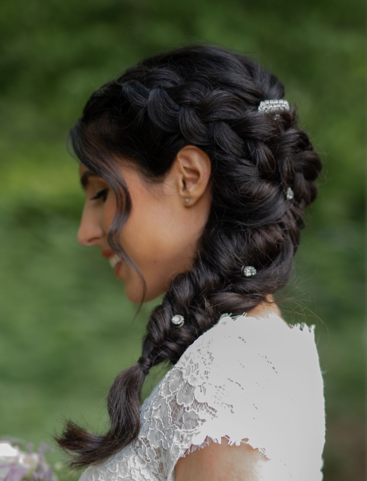 Bride with dark braided hair adorned with crystal pins smiles softly wearing a white lace wedding dress.